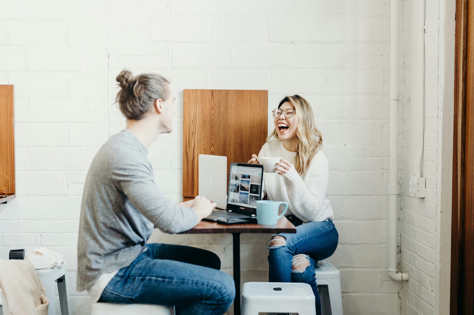 Duas pessoas conversam e riem diante de um notebook em uma mesa com xícaras de café.