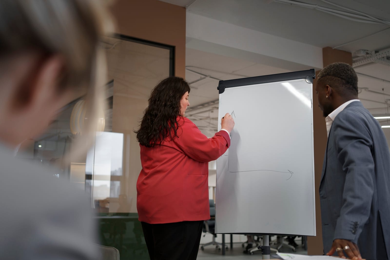 Mulher de blazer vermelho desenha um diagrama em um flipchart enquanto um colega de terno acompanha.