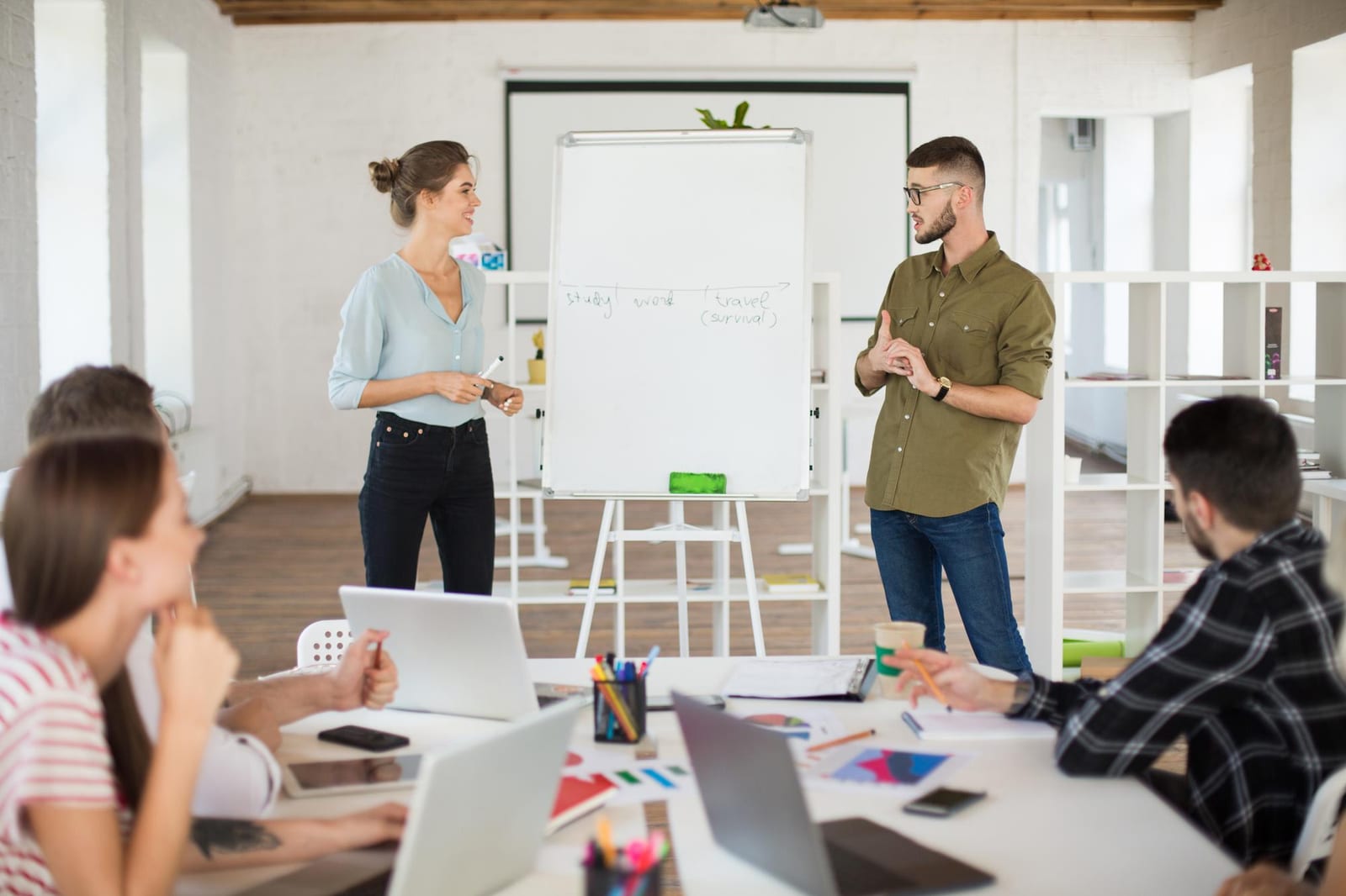 Dois facilitadores conduzem uma dinâmica diante de um flipchart para participantes sentados ao redor de uma mesa com notebooks.